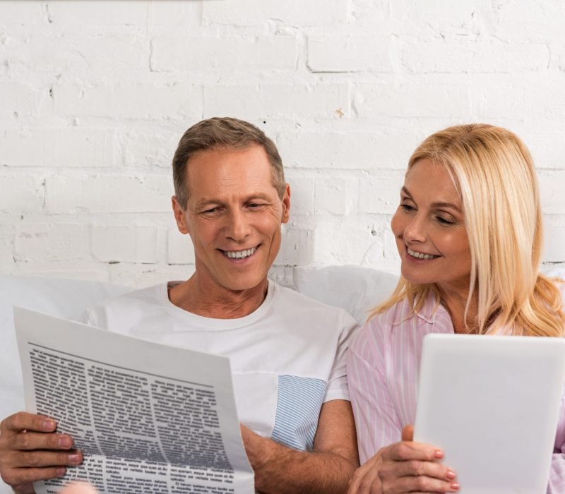 Man reading newspaper to wife with digital tablet on bed